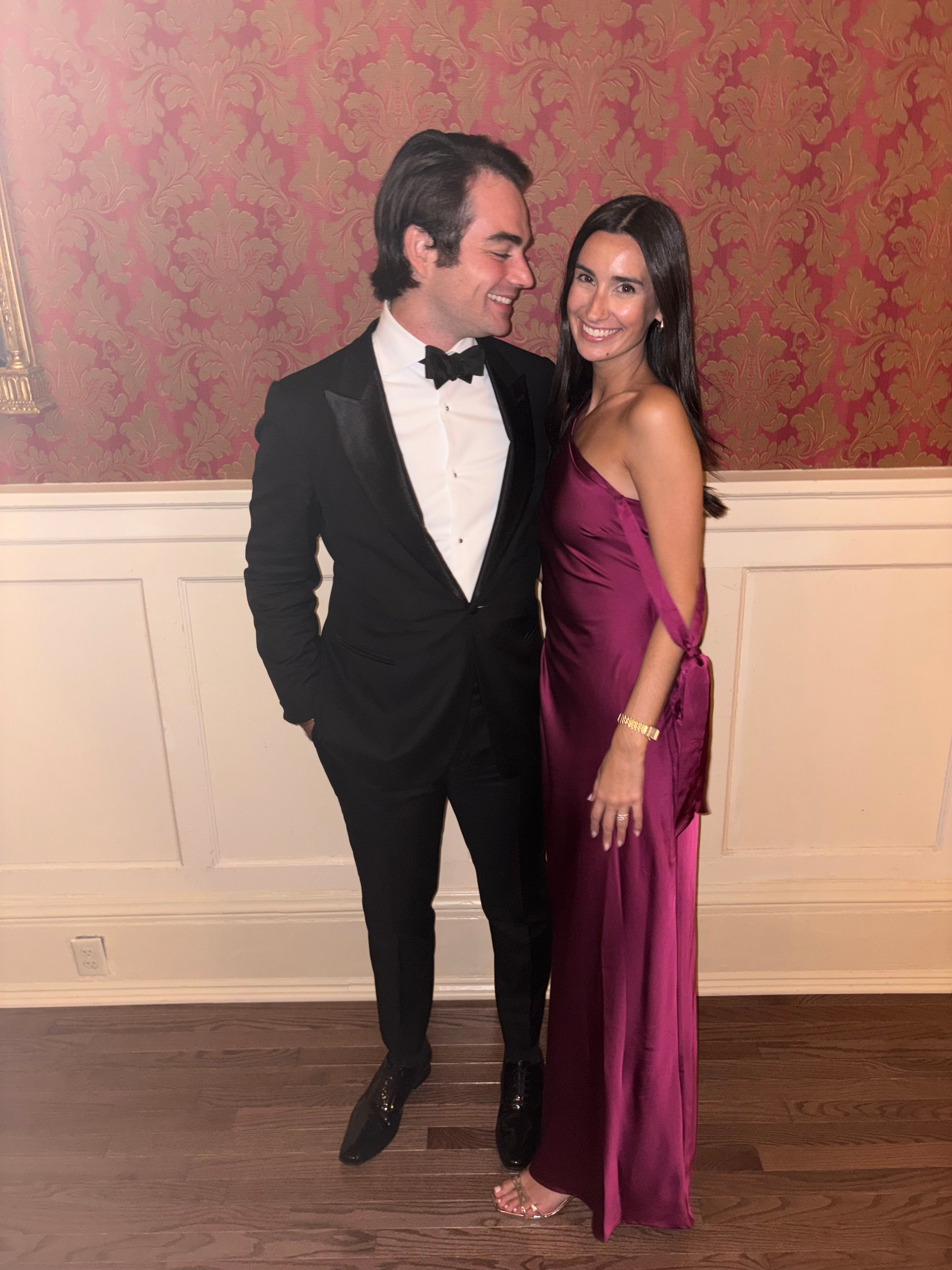 Billy in a tuxedo and Iz in one-shoulder magenta satin, smiling toward the camera against red damask wallpaper and white wainscoting