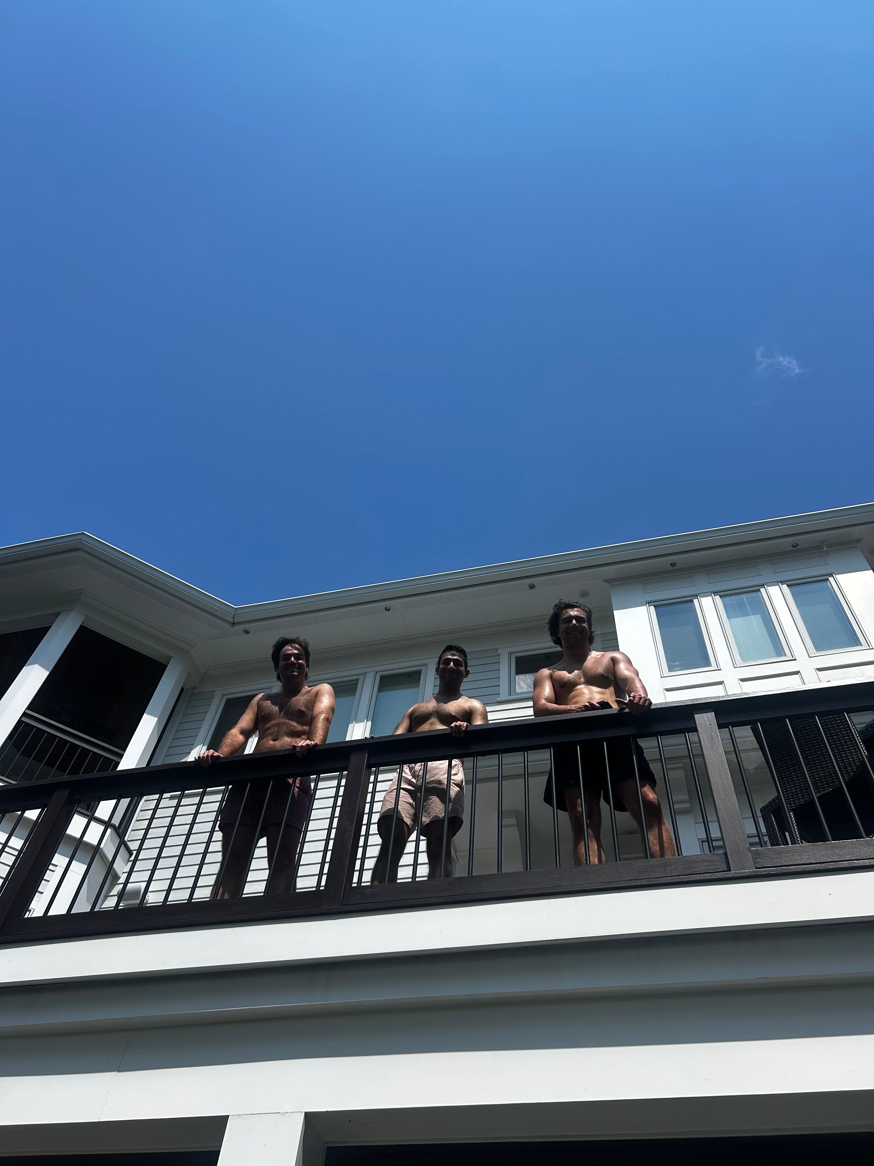 Three people on a balcony looking down, white house and blue sky